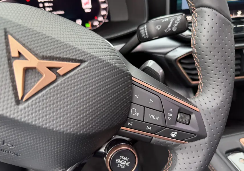 Gros plan sur le volant perforé noir avec surpiqûres cuivrées et logo CUPRA, boutons multifonctions et bouton start/stop.