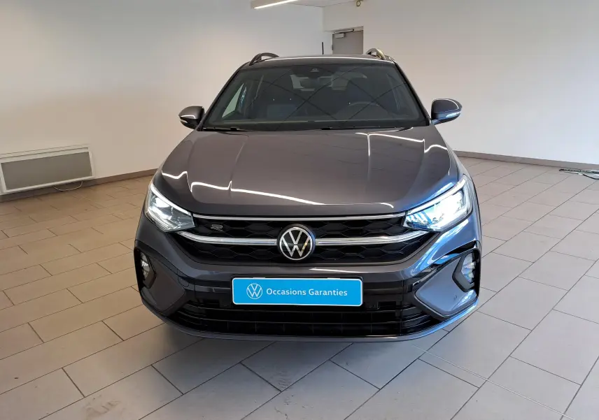 Vue avant d'un Volkswagen Taigo gris cendré 2025 avec calandre R-Line et phares allumés en intérieur showroom.