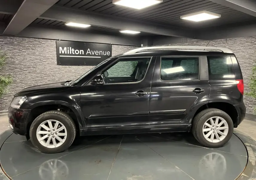 Profil côté gauche d'un Skoda Yeti noir 2015 avec jantes alliage 16'' et barres de toit argentées en intérieur showroom.