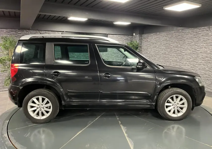 Profil côté gauche du Skoda Yeti noir 2015 avec jantes alliage 16 pouces et toit argenté en intérieur showroom.