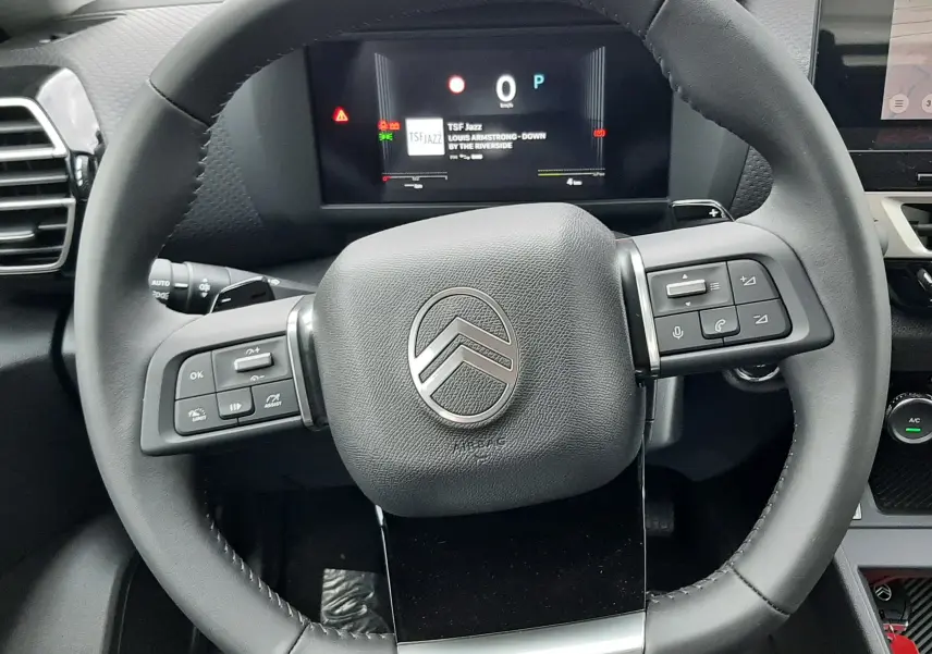 Vue rapprochée du volant multifonction noir du Citroën C4 1.2 Hybride 145ch avec tableau de bord numérique allumé.