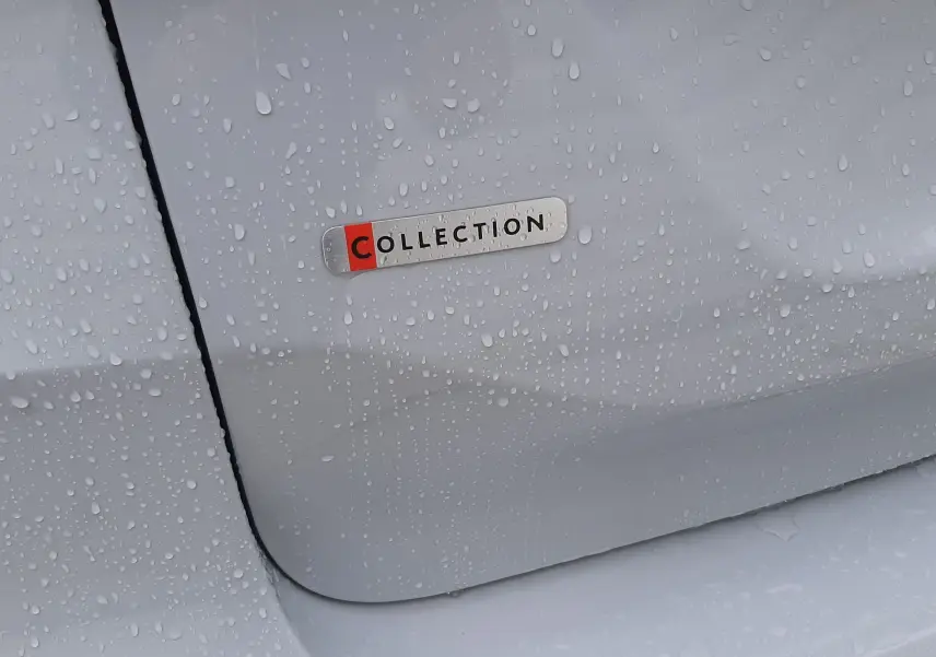 Gros plan sur l'écusson Collection sur la porte arrière côté gauche d'une Citroën C4 blanc Okenite avec gouttes d'eau.