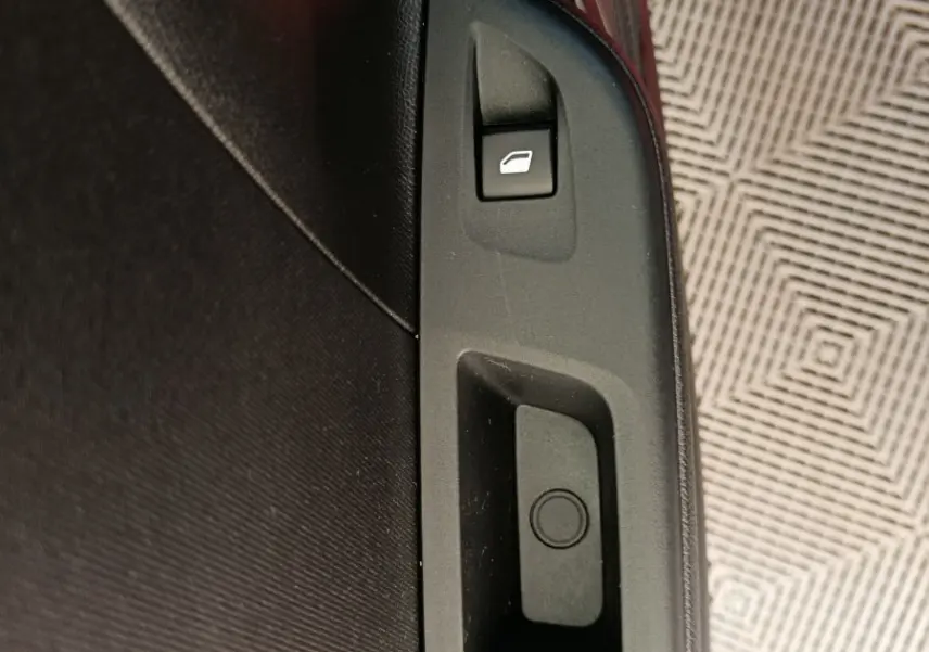 Détail de la commande de lève-vitre sur la porte intérieure noire d’une Opel Corsa rouge visible en arrière-plan.