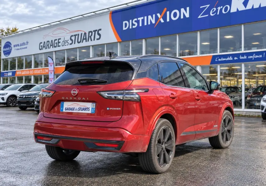 Vue 3/4 arrière d’un Nissan Qashqai 2024 rouge foncé e-Power Tekna garé devant un garage avec toit noir et jantes noires.