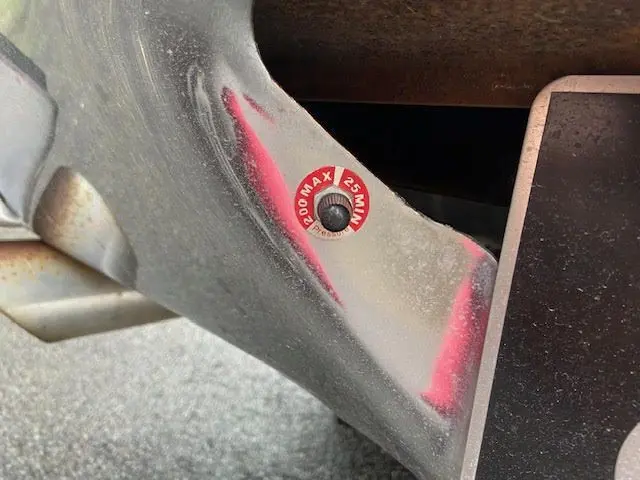 Gros plan sur un détail mécanique avec une étiquette rouge indiquant "200 MAX 25 MIN" sous le châssis d'un Ford Aerostar bordeaux.