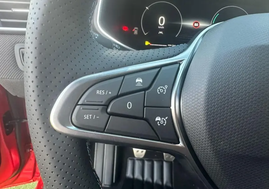 Gros plan sur les commandes au volant perforé de la Renault Clio V rouge flamme, affichage numérique en arrière-plan.