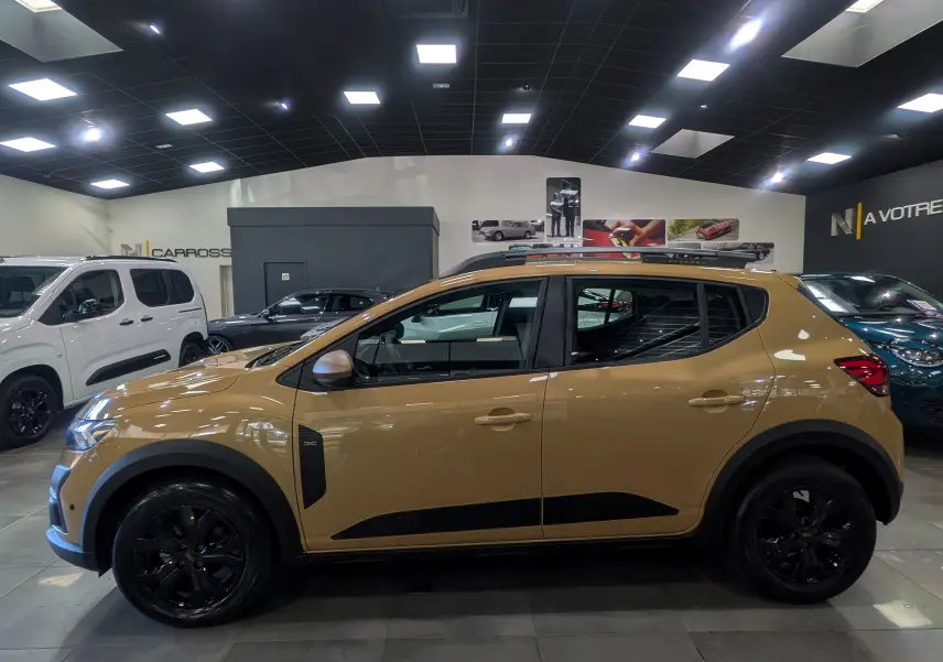 Vue latérale droite d'une Dacia Sandero Stepway beige Safari 2025 avec jantes noires et barres de toit en showroom.
