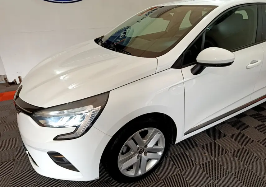 Vue 3/4 avant droit d'une Renault Clio Business Blue dCi 85 blanche avec phares LED allumés en intérieur showroom.