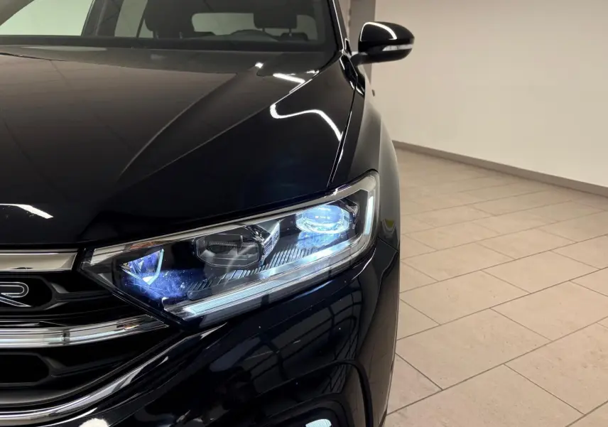Gros plan sur le phare avant droit allumé du Volkswagen T-Roc noir intense, mettant en valeur la calandre R-Line.