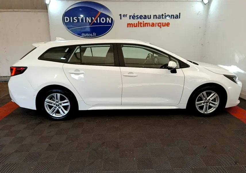 Profil latéral droit d'une Toyota Corolla hybride blanche 2022 en break, stationnée en intérieur sur sol carrelé.