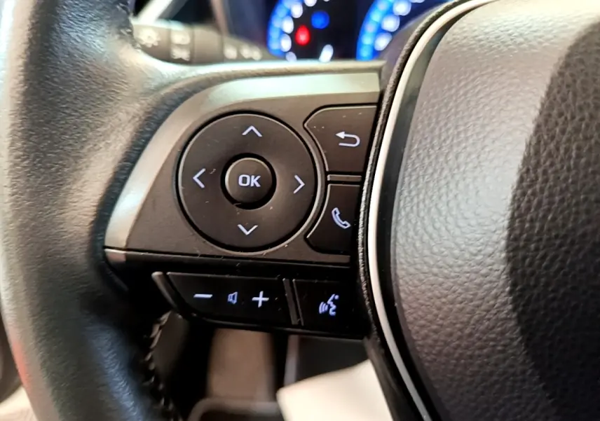 Gros plan sur les commandes noires du volant cuir de la Toyota Corolla hybride blanche, avec tableau de bord lumineux en arrière-plan.