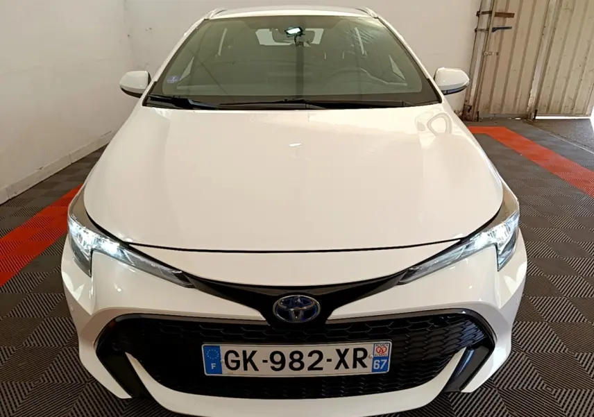 Vue avant d'une Toyota Corolla hybride blanche avec calandre noire et phares LED allumés dans un garage.
