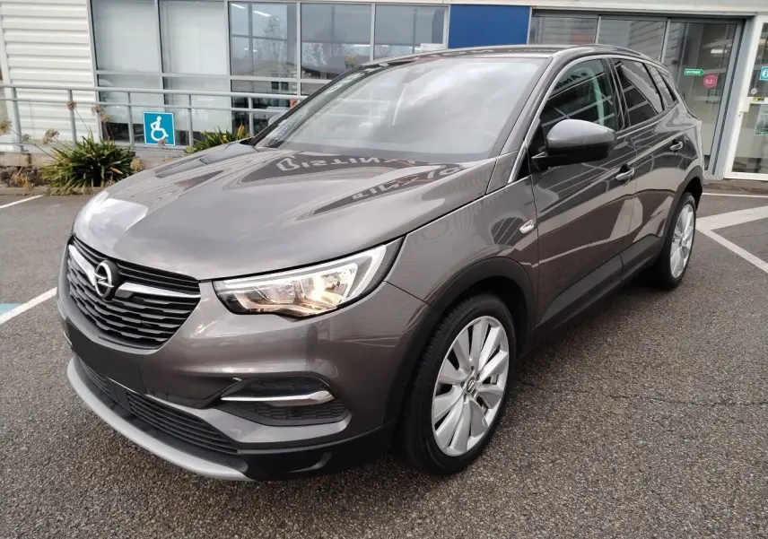 Opel Grandland X gris platinium vu de 3/4 avant droit avec phares allumés sur parking devant bâtiment.
