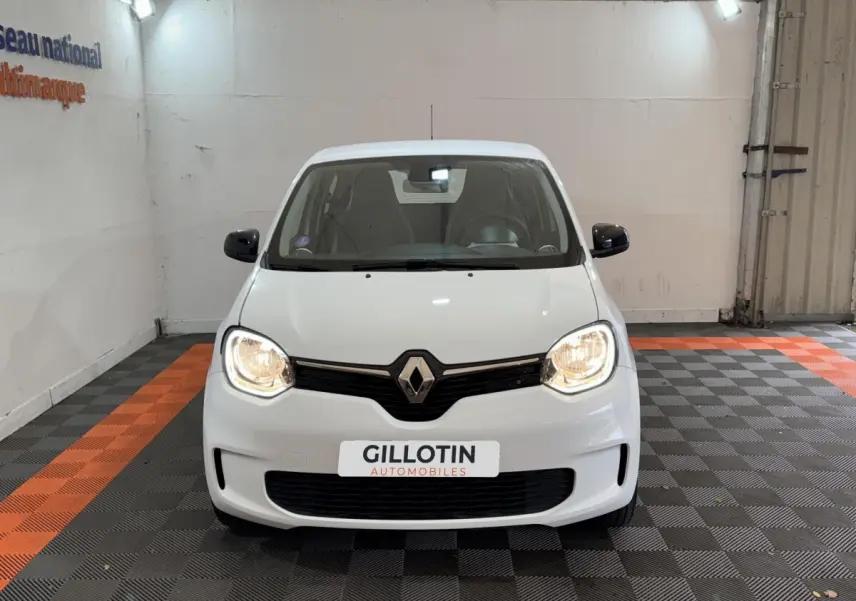 Renault Twingo blanc vue de face avec phares allumés dans un garage au sol gris et orange.