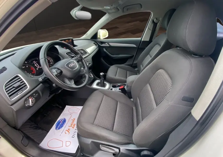 Vue intérieure côté conducteur de l'Audi Q3 2014 avec sièges tissu noir et volant cuir, tableau de bord et levier de vitesse manuelle visibles.