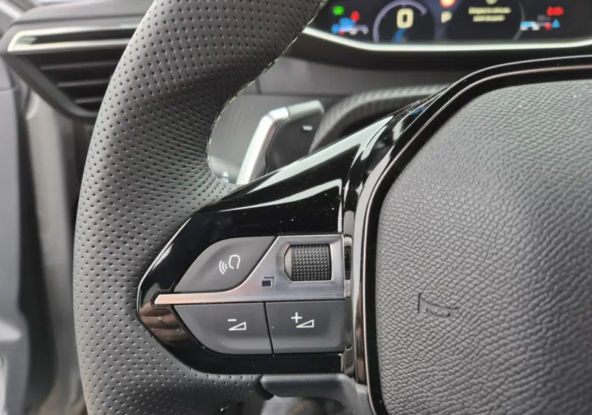 Gros plan sur le volant perforé noir de la Peugeot 208 Hybrid 110 e-DCS6 GT avec commandes intégrées et cockpit numérique en arrière-plan.