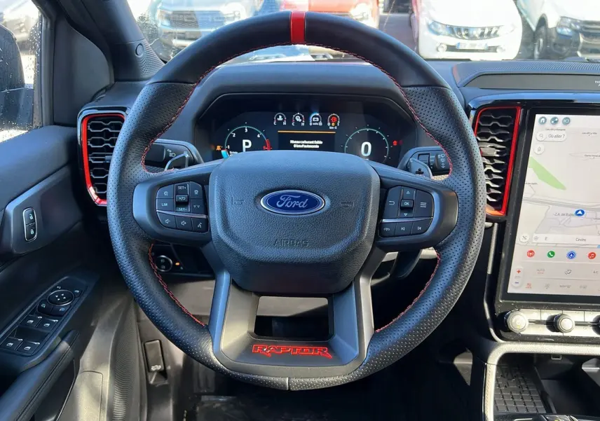 Vue intérieure centrée sur le volant cuir noir avec logo Ford et inscription rouge Raptor, tableau de bord digital et écran tactile.