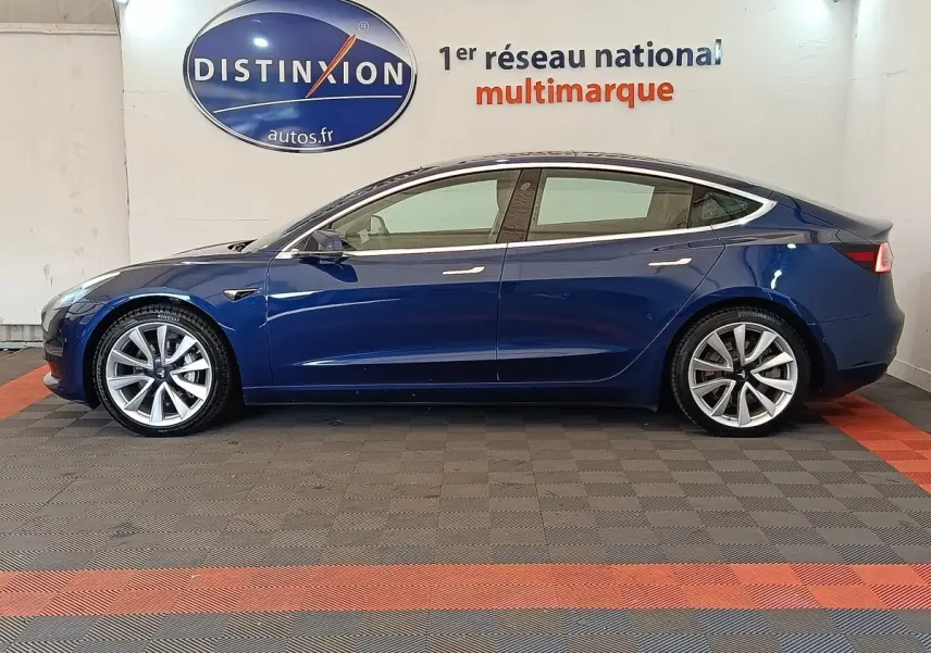 Profil droit d'une Tesla Model 3 Long Range bleu électrique, avec jantes argentées et lignes épurées visibles en intérieur showroom.