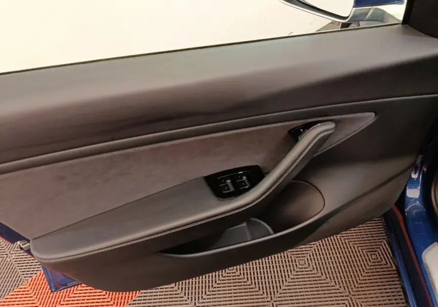 Vue intérieure sur la porte côté gauche d’une Tesla Model 3 bleu, avec commandes de vitres et poignée intégrée.