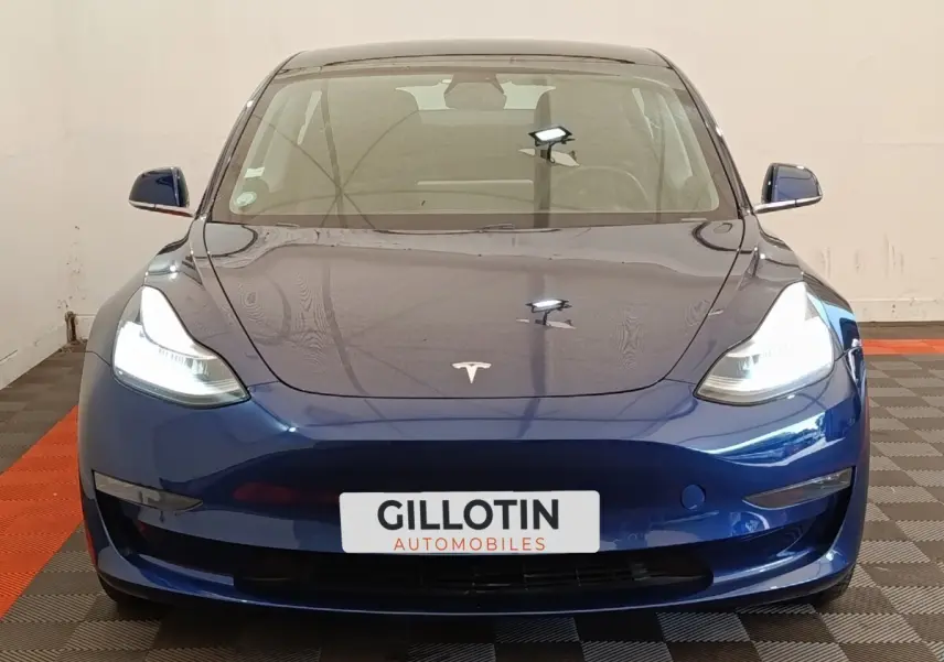 Vue frontale d'une Tesla Model 3 Long Range AWD bleu électrique avec phares allumés en intérieur.