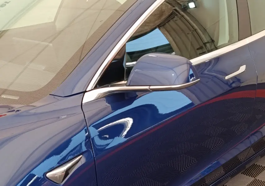 Détail du côté gauche bleu brillant d'une Tesla Model 3 2020 avec rétroviseur et poignée de porte visibles.