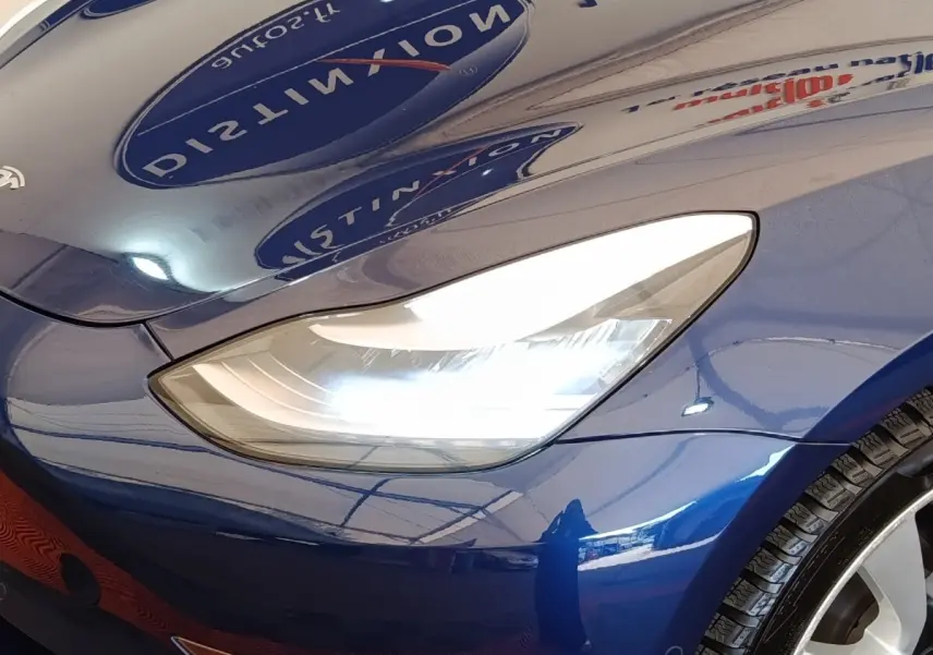 Gros plan sur le phare avant droit d’une Tesla Model 3 Long Range bleu électrique, avec reflet brillant sur la carrosserie.