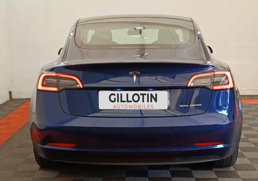 Vue arrière d'une Tesla Model 3 Long Range bleu électrique avec feux LED allumés et logo central visible.