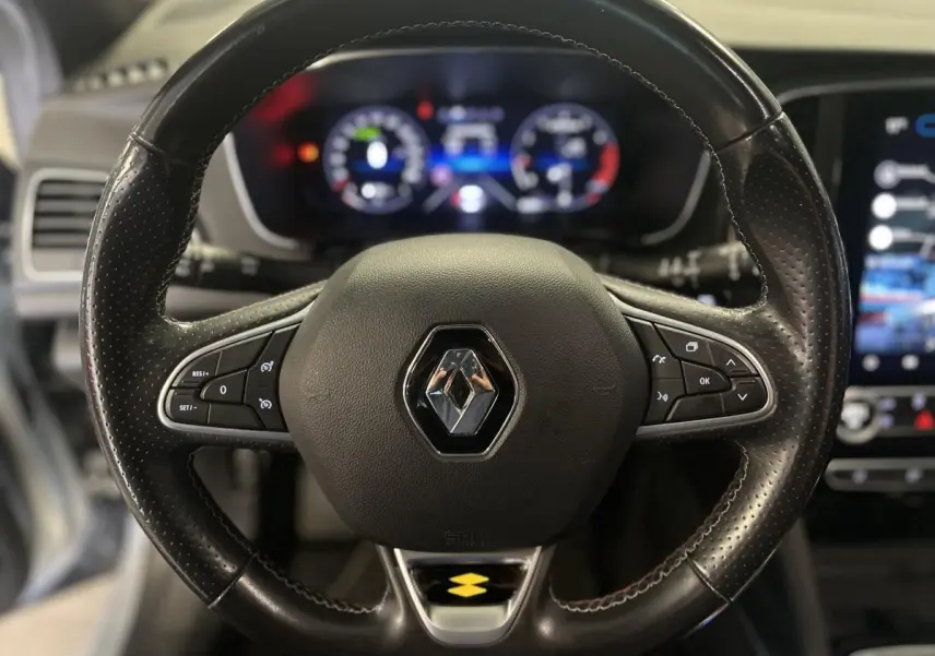 Gros plan sur le volant cuir noir R.S. Line avec badge Renault, tableau de bord numérique flou en arrière-plan.
