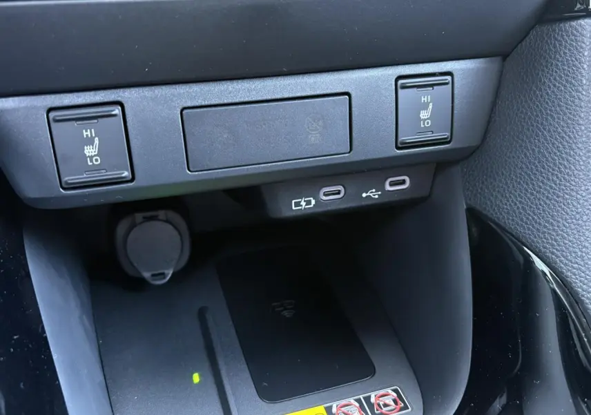 Gros plan sur la console centrale de la Toyota Yaris hybride 2025 montrant les commandes des sièges chauffants et un chargeur à induction noir.