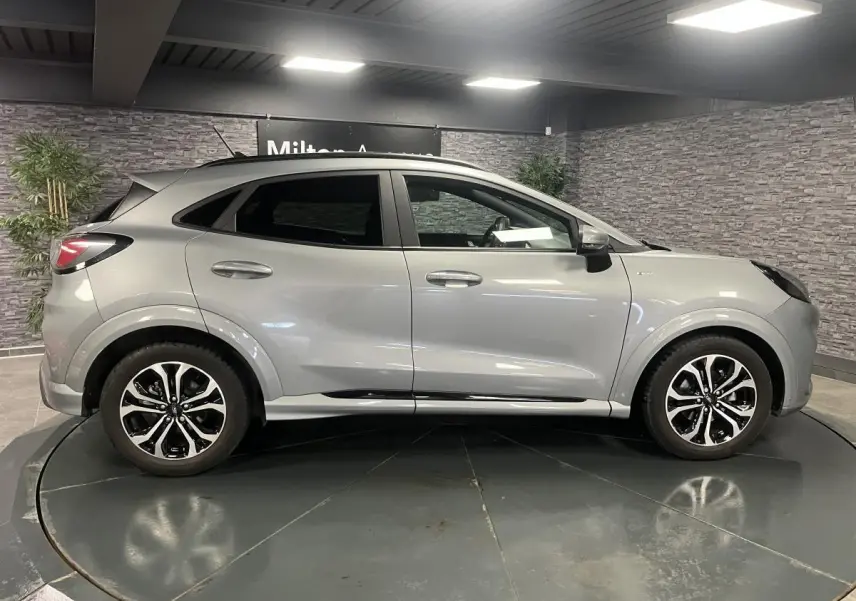 Ford Puma gris Solar en profil droit, avec jantes alliage noires et barres de toit noires visibles en intérieur showroom.