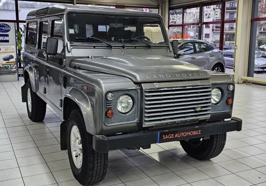Vue 3/4 avant d'un Land Rover Defender 110 Station Wagon S gris, avec calandre distinctive et phare rond en intérieur showroom.