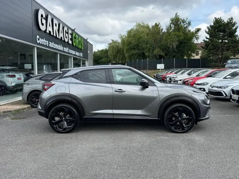 Profil droit d'un Nissan Juke 2025 gris métallisé avec jantes noires, garé devant un garage automobile.