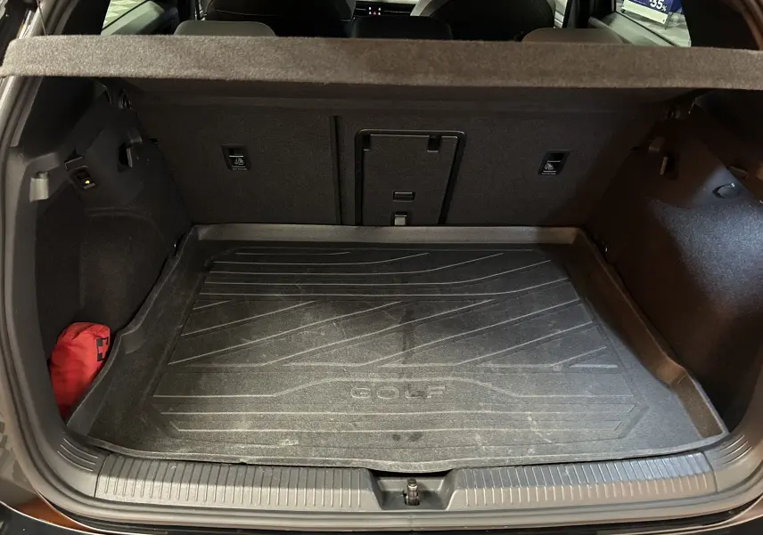 Coffre ouvert vu de l'arrière d'une Volkswagen Golf VIII eTSI noir métal, avec tapis de coffre marqué Golf et trousse de secours rouge.
