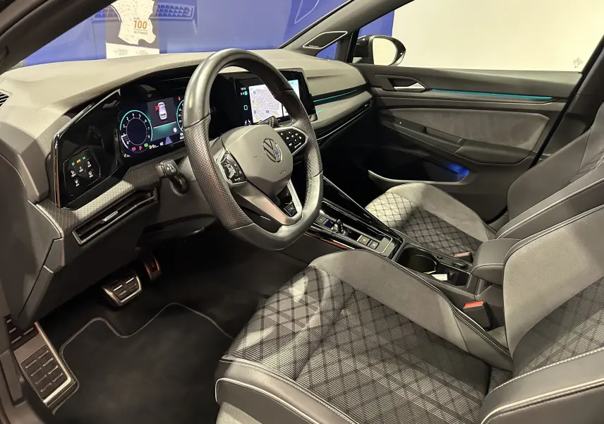 Vue intérieure côté conducteur de la Volkswagen Golf VIII eTSI 150CH R-Line noire, avec tableau de bord digital et sièges en tissu gris à motifs.