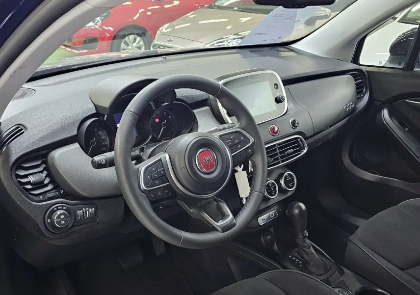 Intérieur de la FIAT 500X 2024 en bleu, vue côté conducteur montrant le volant, tableau de bord et console centrale.