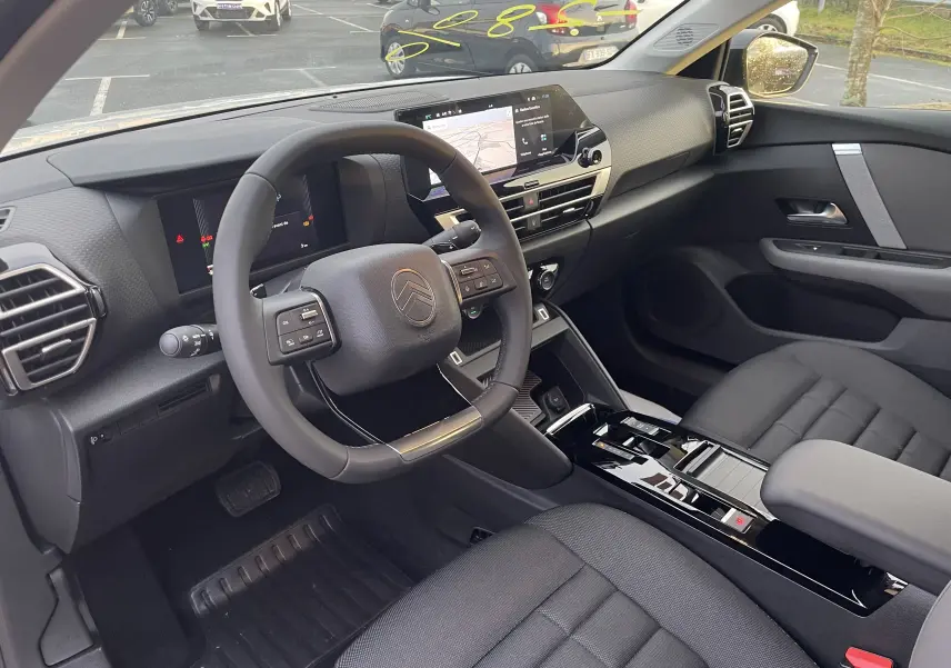 Vue intérieure côté conducteur de la Citroën C4 1.2 Hybride 145ch 2025 avec tableau de bord numérique et sellerie tissu noir.