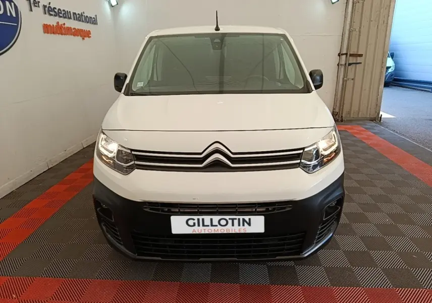 Vue frontale d’un Citroën Berlingo blanc avec pare-chocs noir et phares allumés en intérieur showroom.