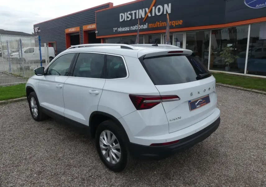 Vue 3/4 arrière droite d'un SUV SKODA KAROQ blanc garé devant un bâtiment commercial avec vitres arrière teintées.
