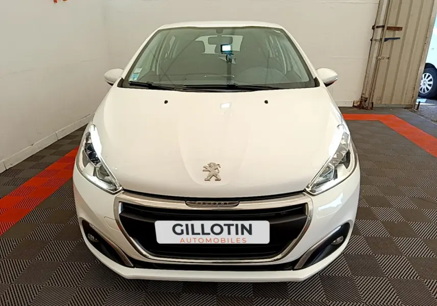 Vue avant d'une Peugeot 208 Puretech 82 Signature blanche, avec calandre chromée et phares allumés en intérieur showroom.