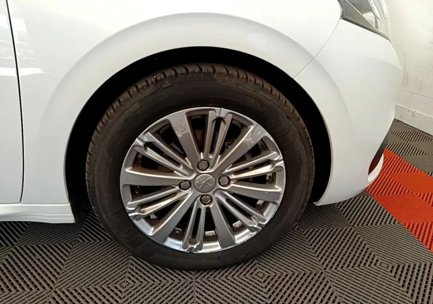 Gros plan sur la roue avant gauche d'une Peugeot 208 blanche, mettant en valeur la jante alliage au design moderne.