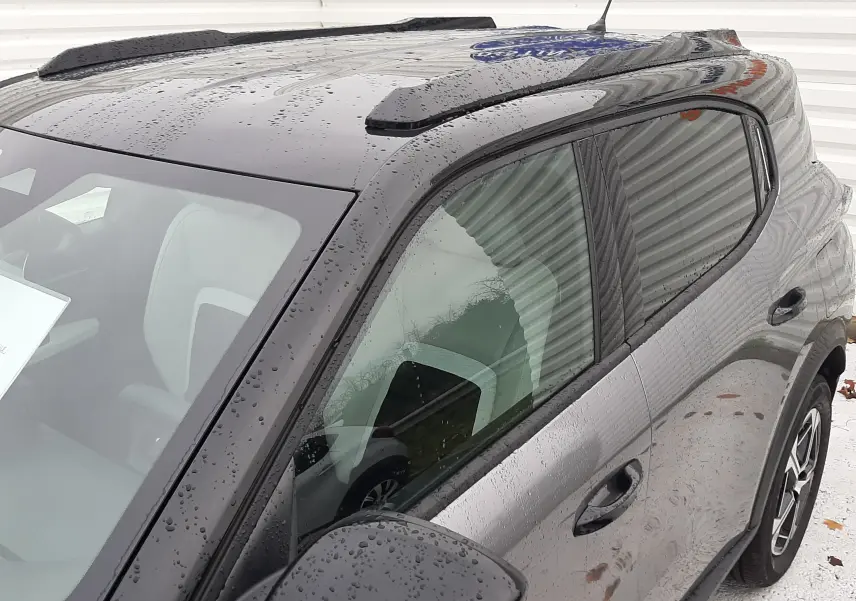 Vue 3/4 avant droit du Citroën C3 Aircross gris avec toit noir brillant et barres de toit noires sous la pluie.