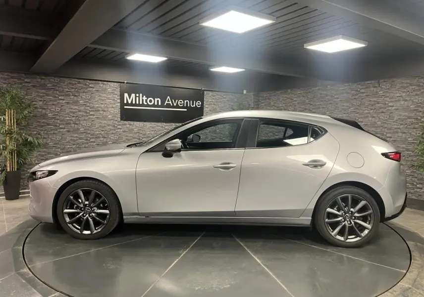 Profil côté gauche d'une Mazda3 gris métallisé 2023 avec jantes alliage 18 pouces dans un showroom.