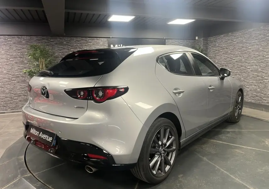 Vue 3/4 arrière droite de la Mazda3 gris métallisé avec toit noir et jantes alliage 18 pouces dans un showroom.
