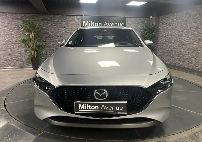 Vue frontale d'une Mazda3 gris métallisé avec calandre noire et éclairage LED, en intérieur showroom.