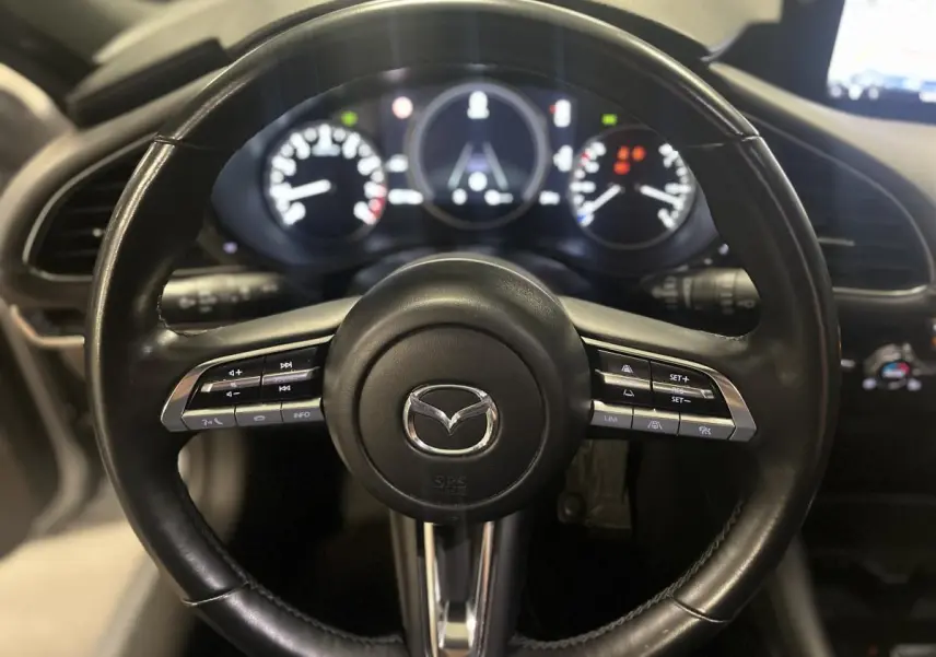 Volant cuir noir de Mazda3 2023 vu de face, avec commandes intégrées et tableau de bord numérique en arrière-plan.