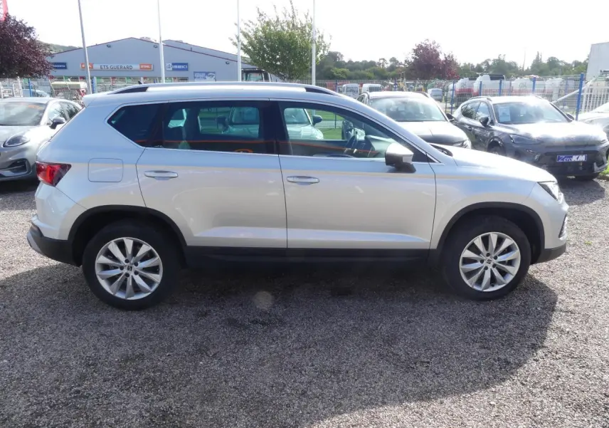 Vue de profil côté gauche d'un SEAT Ateca gris clair 2023 avec jantes alliage 17 pouces et barres de toit chromées.