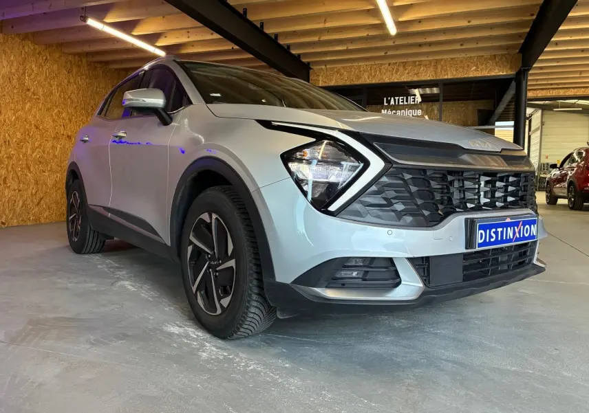KIA Sportage hybride gris en 3/4 avant droit, avec calandre noire et phares anguleux, dans un garage éclairé.