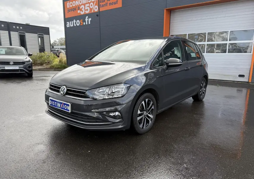 Volkswagen Golf Sportsvan gris Urano vue 3/4 avant droit sur parking devant un bâtiment commercial.