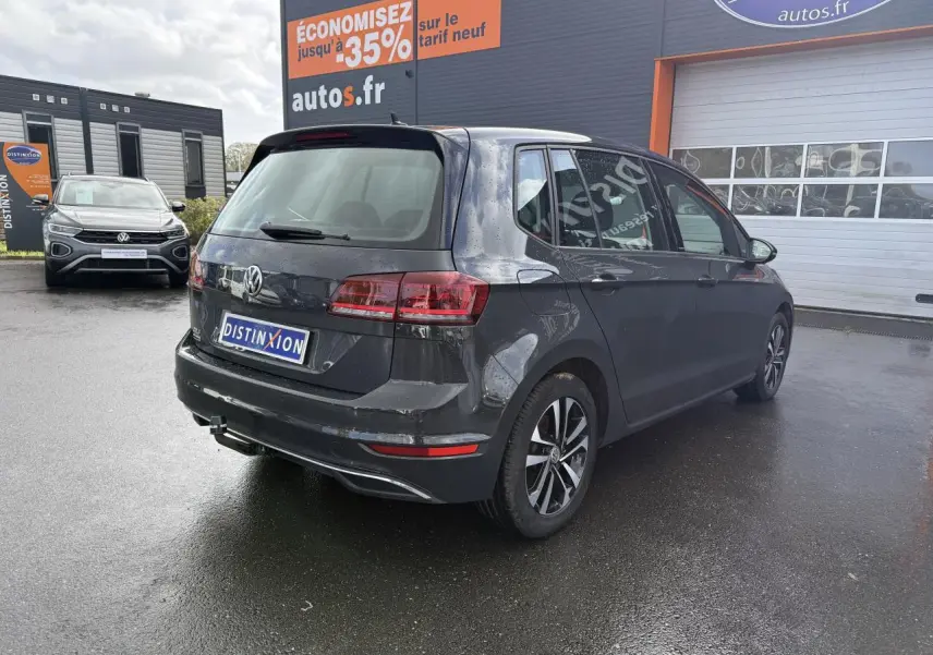 Vue 3/4 arrière droite d'une Volkswagen Golf Sportsvan gris Urano stationnée devant un garage autos.fr.