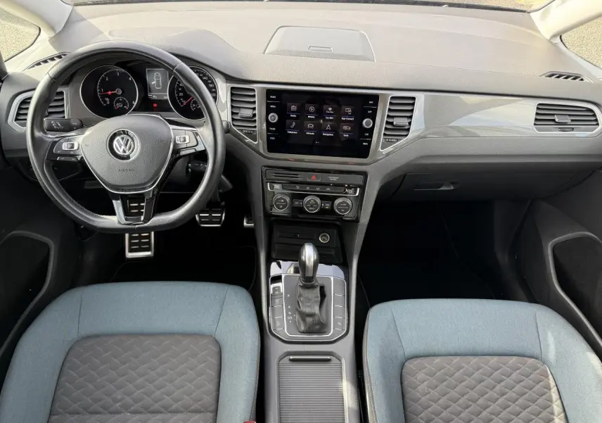 Vue intérieure avant du Volkswagen Golf Sportsvan 2019, tableau de bord noir avec écran tactile et sièges bicolores gris et bleu.